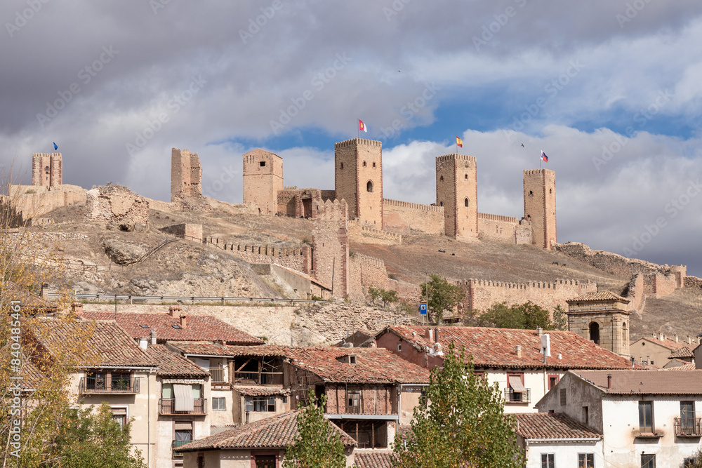 Ancient stone castle, rustic village below, flags on towers, partly cloudy sky, historical ambiance, morning or afternoon light Ancient stone castle, rustic village below, flags on towers, partly cloudy sky, historical ambiance, morning or afternoon light