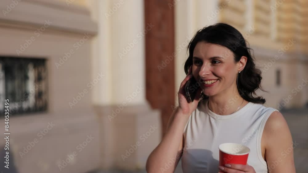 Woman talking, Coffee drinking, Fashion stroll. In white top and jeans, young woman seen walking through city, talking on phone and holding red coffee cup. Woman talking, Coffee drinking, Fashion stroll. In white top and jeans, young woman seen walking through city, talking on phone and holding red coffee cup.