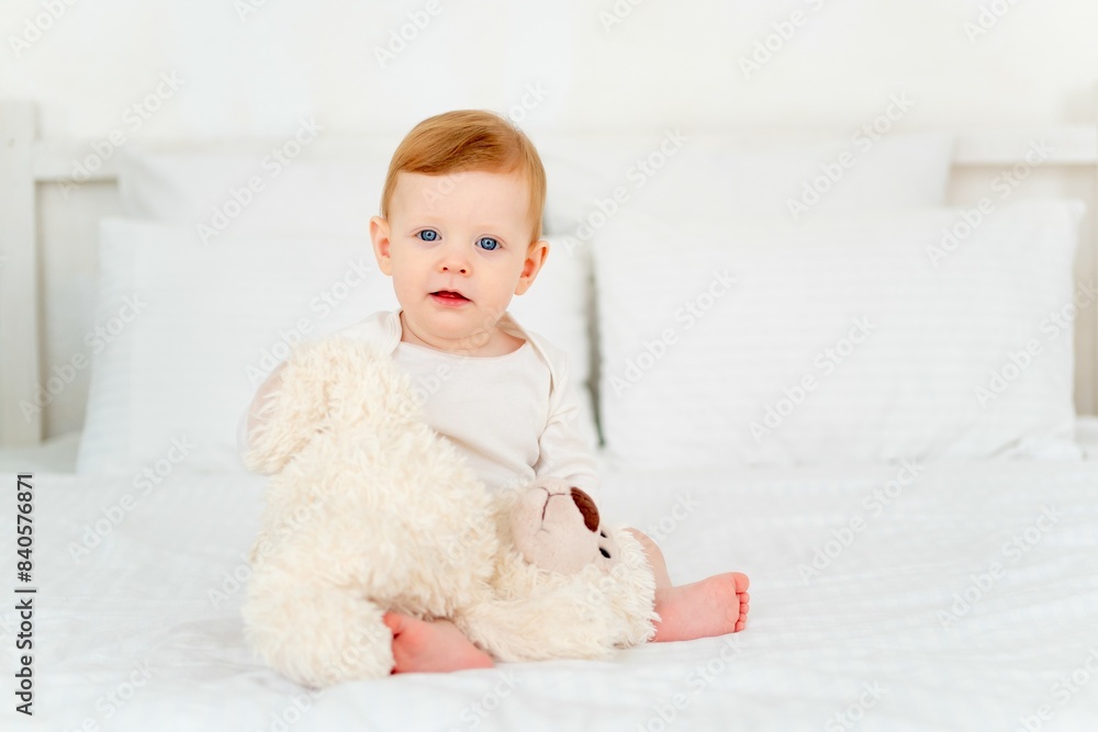 A cute little smiling baby boy in a white bodysuit is sitting on a bed at home in a bright bedroom with a soft teddy bear, a six-month-old baby with blue eyes is smiling or laughing A cute little smiling baby boy in a white bodysuit is sitting on a bed at home in a bright bedroom with a soft teddy bear, a six-month-old baby with blue eyes is smiling or laughing