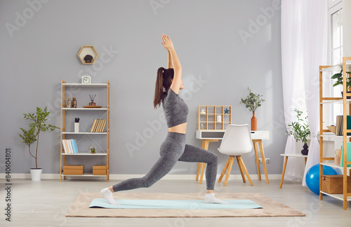 Side view of a beautiful young woman in sports clothes having an indoor yoga workout at home, standing on an exercise mat in the living room interior and doing a Crescent lunge aka Ashta Chandrasana photo