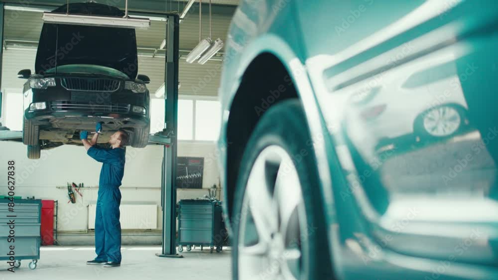 Camera view of Caucasian serviceman working on broken car in background. Hardworking male standing under car which located on two-post lift. Actively fixing detail or bolt with wrench. Camera view of Caucasian serviceman working on broken car in background. Hardworking male standing under car which located on two-post lift. Actively fixing detail or bolt with wrench.
