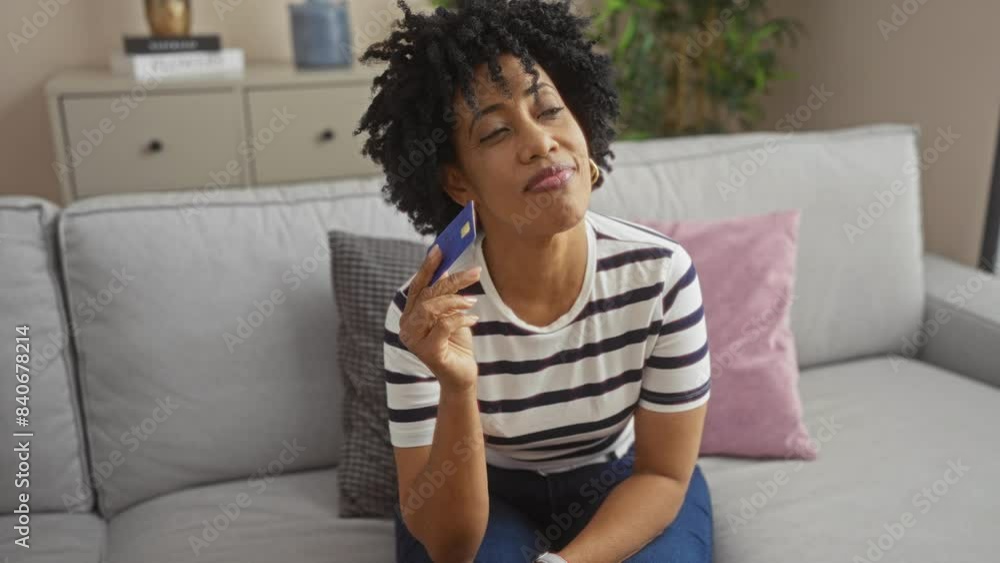 A young african american woman with curly hair pondering while holding a credit card in a modern living room, dressed in casual wear and sitting on a comfortable sofa. A young african american woman with curly hair pondering while holding a credit card in a modern living room, dressed in casual wear and sitting on a comfortable sofa.