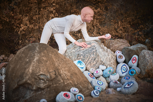 Young hairless girl with alopecia in white futuristic costume pensively examines stone with eye at surreal landscape, symbolizes introspection and reevaluation of individuality photo