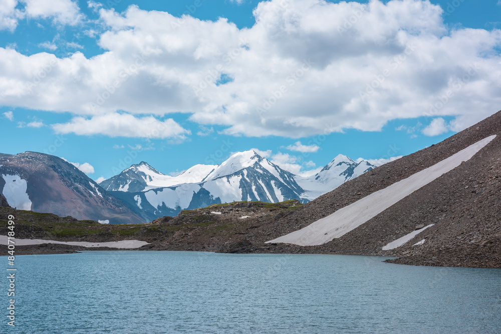 Most beautiful view to azure alpine lake against snow-covered range with few pointy peaks. Ripples on turquoise water of mountain lake against three snowy peaked tops under clouds in cloudy blue sky.