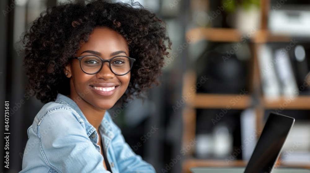 A young woman wearing glasses smiles warmly as she works on a laptop in a modern office setting A young woman wearing glasses smiles warmly as she works on a laptop in a modern office setting