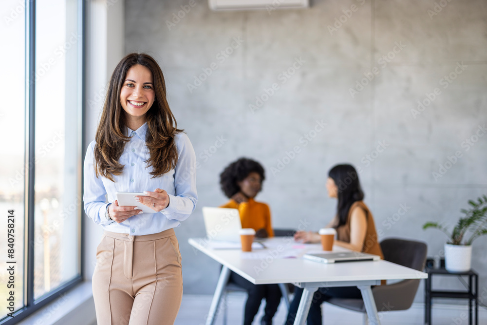 A cheerful Caucasian businesswoman explores a digital tablet in a sunlit modern office, with colleagues engaged in work behind her.