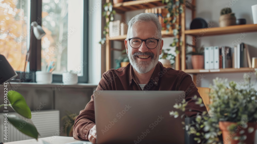 A smiling man sits comfortably at his desk, working on his laptop, with a bright and airy home office filled with plants and books in the background. A smiling man sits comfortably at his desk, working on his laptop, with a bright and airy home office filled with plants and books in the background.