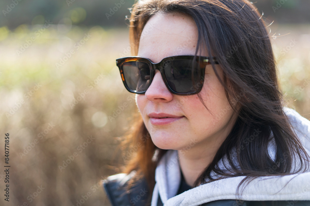 A cute dark-haired girl in sunglasses, a vest and a hoodie stands smiling in a spring park A cute dark-haired girl in sunglasses, a vest and a hoodie stands smiling in a spring park