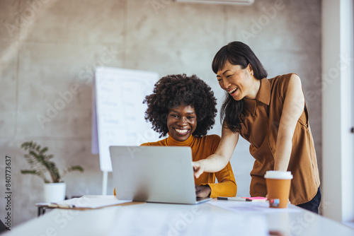 An Asian and a Black businesswoman collaborate joyfully over a laptop in a modern office setting, dressed smartly, illustrating teamwork and professional partnership.