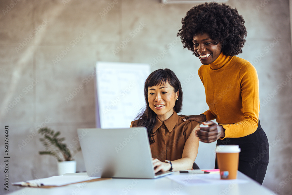 An Asian and a Black businesswoman engage in a collaborative effort, reviewing content on a laptop in a well-lit workspace, dressed in smart casual attire. An Asian and a Black businesswoman engage in a collaborative effort, reviewing content on a laptop in a well-lit workspace, dressed in smart casual attire.