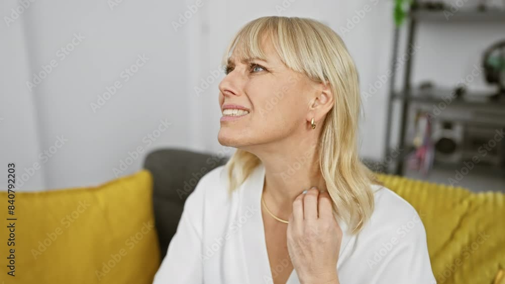A blonde woman experiencing discomfort touches her temple while seated on a couch indoors. A blonde woman experiencing discomfort touches her temple while seated on a couch indoors.