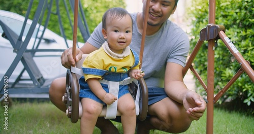 A joyful moment of a father pushing his smiling baby in a swing at a garden playground. The baby, dressed in a yellow shirt and denim overalls, is safely strapped in, enjoying the outdoor playtime.