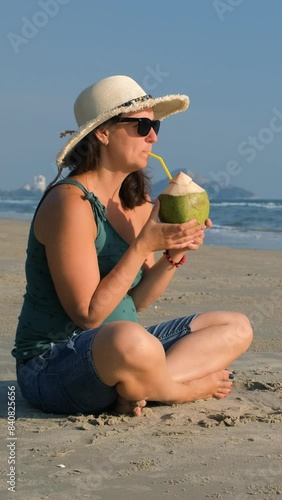 Vertical video. Cheerful young woman in straw hat enjoying summer vacation and drinking coconut water on the beach