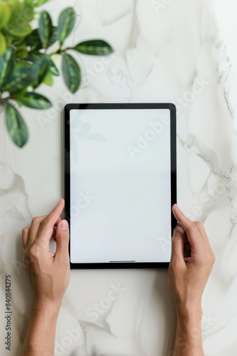 Top View of Hands Holding a Tablet With Blank Screen on a White Table Surrounded by Greenery