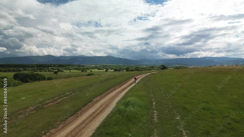 Drone following woman cyclist riding bike in incredible epic landscape surrounded by mountains and green fields. Gravel cycling on dirt unpaved road in natural park. Gravel cycling concept. Drone following woman cyclist riding bike in incredible epic landscape surrounded by mountains and green fields. Gravel cycling on dirt unpaved road in natural park. Gravel cycling concept.