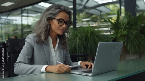 SProductive Business Woman in Green Office. A focused professional woman jotting down notes beside a laptop in a bright  plant-filled office space.