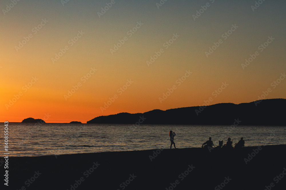 Obraz premium While watching the evening sunset on the shores of Agawa Bay campground a silhouetted young woman takes a photograph of her family while they all relax along the beach