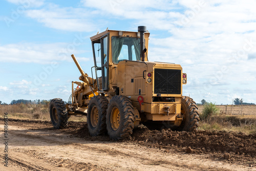 Heavy machinery is working on a dirt road in a rural setting under a partly cloudy sky. The image showcases two yellow graders and a green tractor, emphasizing construction and agricultural themes
