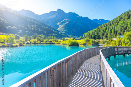 A tranquil wooden boardwalk bridge curves above the vibrant blue waters of Lake in Sulden, Italy. With the majestic Italian Alps towering in the background under a clear azure sky. photo