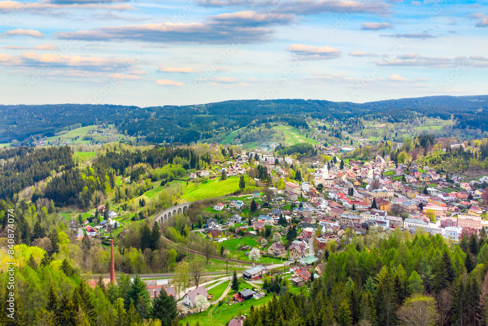 Fototapeta premium A vibrant springtime panorama over Smrzovka town, nestled amidst lush green hills and forests under a partly cloudy sky. Czechia