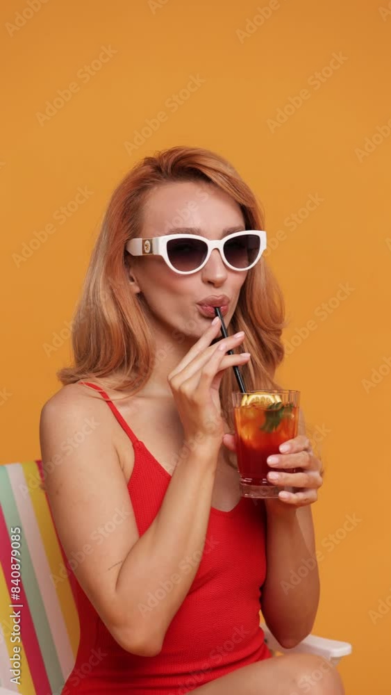 A stylish woman in a red swimsuit and white sunglasses enjoys a refreshing drink against an orange background. Capturing the essence of summer relaxation and fashion. A stylish woman in a red swimsuit and white sunglasses enjoys a refreshing drink against an orange background. Capturing the essence of summer relaxation and fashion.