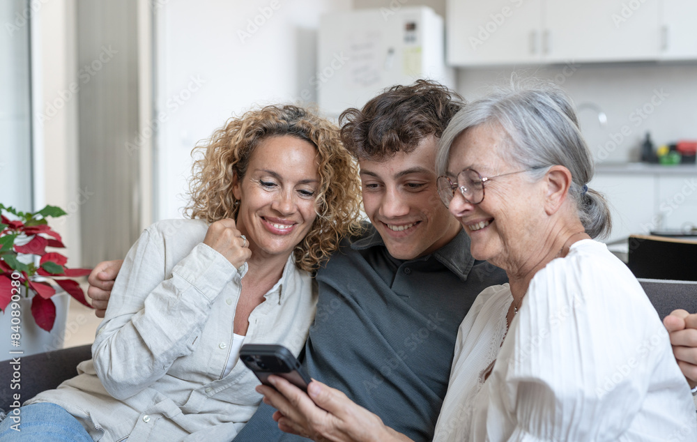 Obraz premium Happy multigenerational family watching funny videos on smartphone together. Grandmother, mature daughter and young grandson having fun and laughing while sitting on the sofa at home