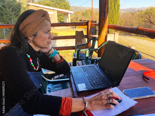 a woman with long hair, wearing a Brown headband and colourful neklace, Works on a laptop while sitting at an outdoor wooden table. she is surrounded by lush greenery and a scenic view of mountain in photo