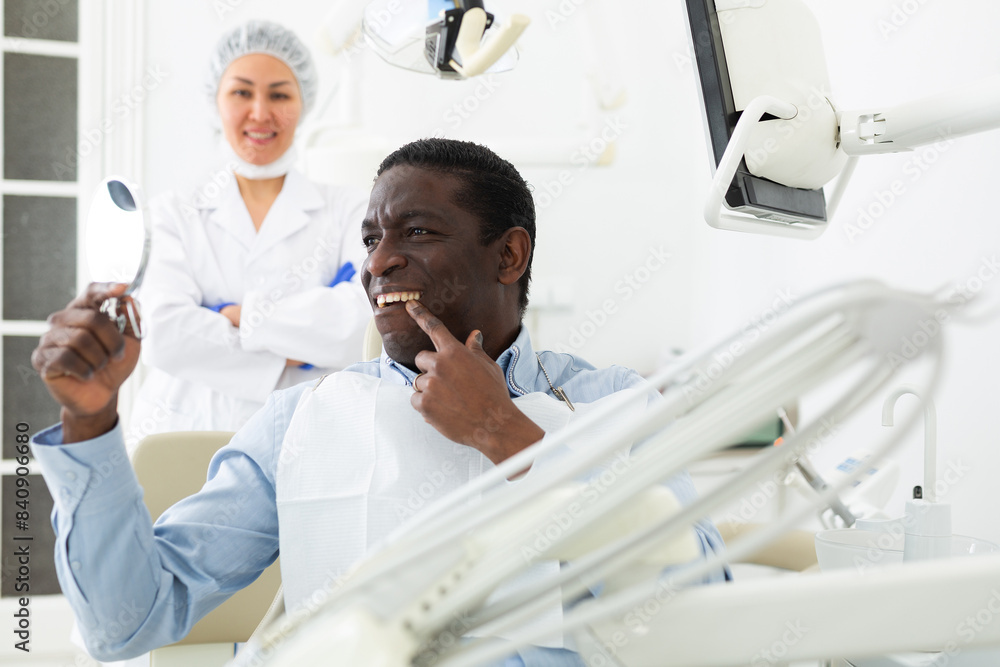 Fototapeta premium African-american man sitting on dental chair and looking at mirror after tooth restoration. Asian woman dentist standing in background.