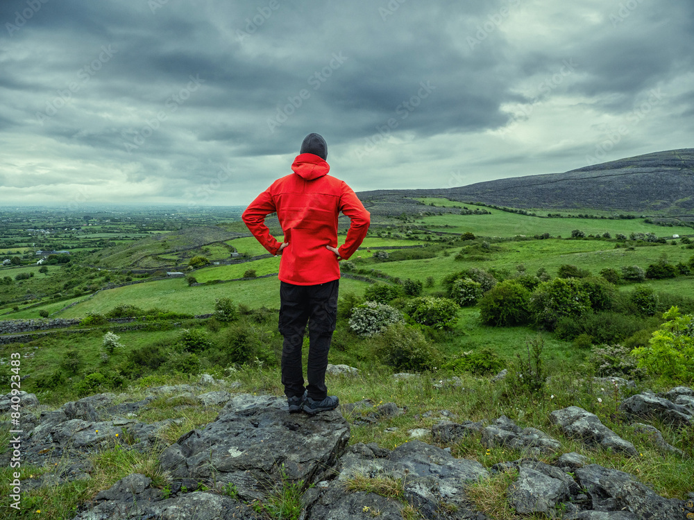 Obraz premium Man in red jacket looking at stunning nature scenery with mountains and dramatic sky. Burren, Ireland. Irish nature scene. Travel and tourism concept. Outdoor activity and sightseeing.