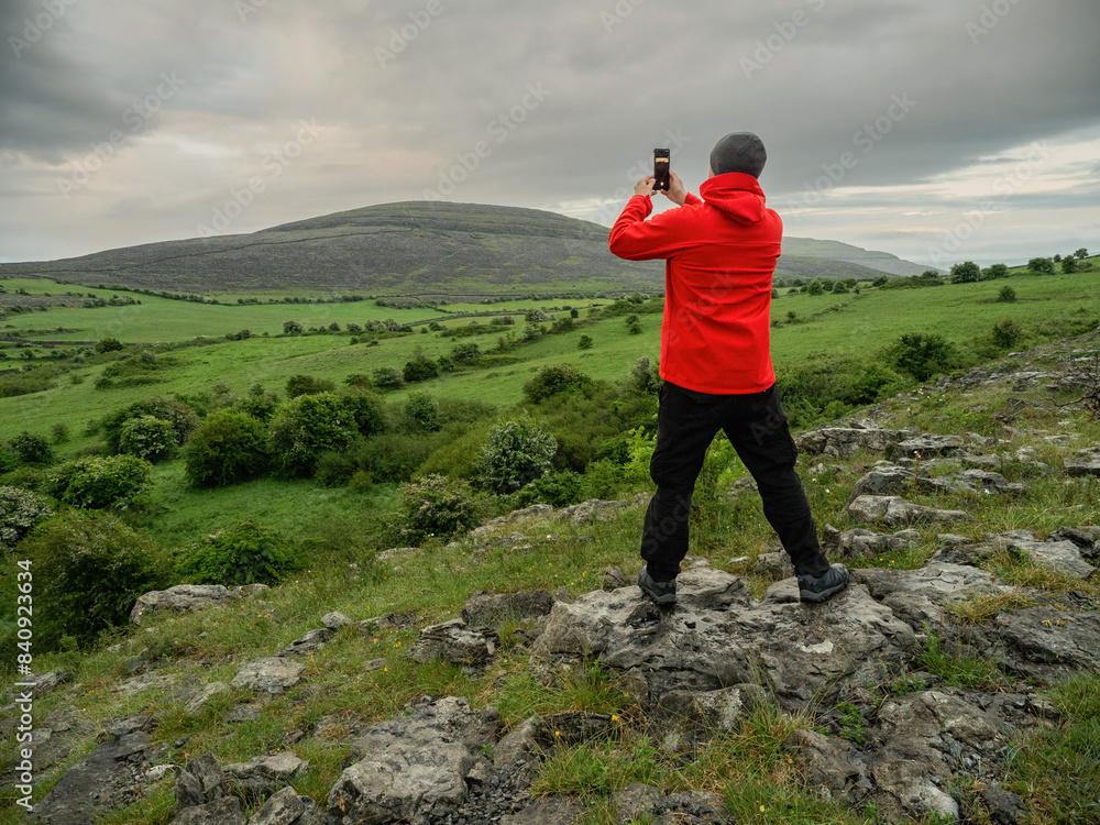 Obraz premium A male tourist in red outdoor jacket taking picture of stunning nature scene on his smart phone. Travel concept. Beautiful nature of Burren with hills and dramatic sky. West of Ireland.