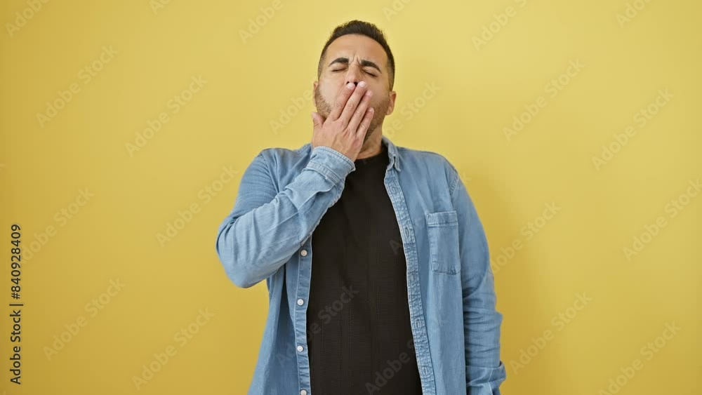 Tired and bored! young hispanic guy in denim shirt, yawning and covering mouth with hand, battles sleepiness - isolated on a yellow background. Tired and bored! young hispanic guy in denim shirt, yawning and covering mouth with hand, battles sleepiness - isolated on a yellow background.