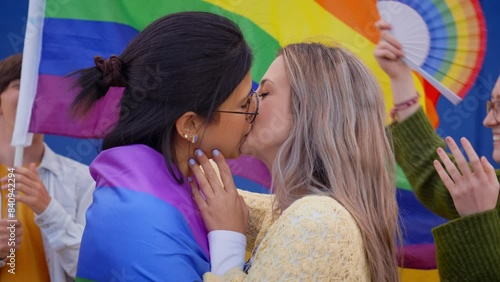 Young lesbian couple kissing on gay pride day outdoors. Group of LGBT community celebrating party on background with rainbow flag and fans. Types of sexuality on generation z photo