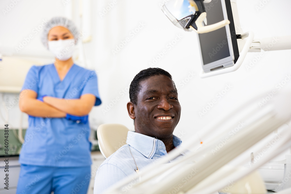 Fototapeta premium African-american man patient sitting on dental chair and smiling. Asian woman dentist standing in background.