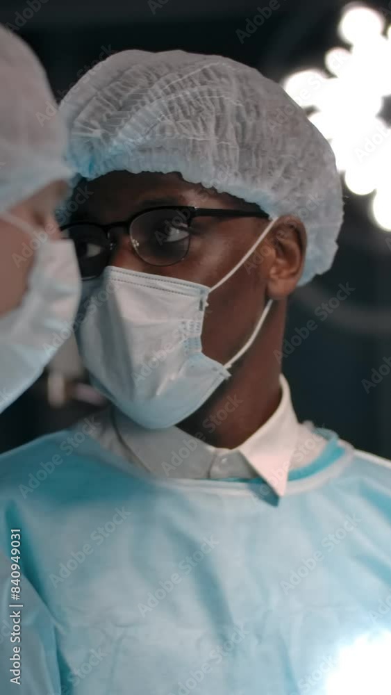 Diverse men in medical masks and hats discussing surgery plan while standing near lamp in operating theater. Vertical shot. Diverse men in medical masks and hats discussing surgery plan while standing near lamp in operating theater. Vertical shot.