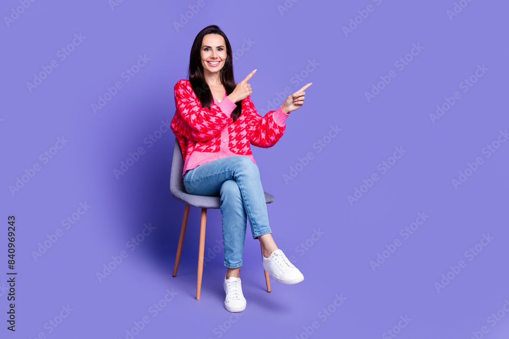 Full length photo of shiny attractive lady dressed pink cardigan sitting chair pointing empty space isolated violet color background Full length photo of shiny attractive lady dressed pink cardigan sitting chair pointing empty space isolated violet color background