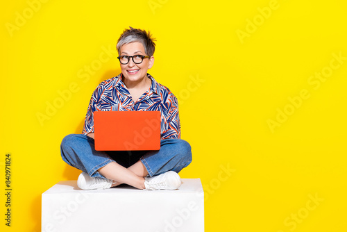 Full body photo of attractive retired woman sit cube hold netbook dressed stylish colorful clothes isolated on yellow color background