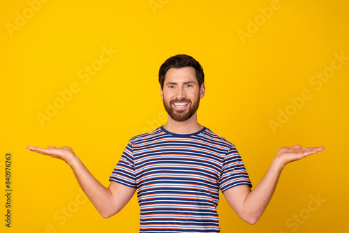 Portrait of pleasant guy with bristle wear striped t-shirt comparing objects on arms empty space isolated on yellow color background