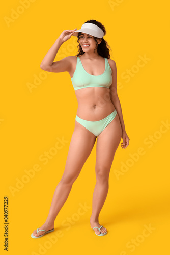 Beautiful young African-American woman in stylish turquoise swimsuit and cap on yellow background