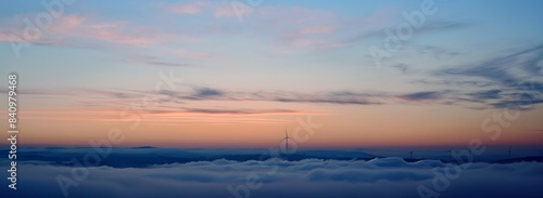 Three wind turbines rise against the backdrop of an orange and blue sky  with white clouds partially hiding them.