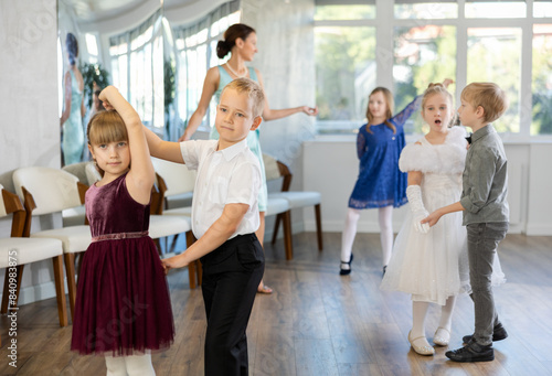 Smiling junior schoolchildren in party dresses learning dancing waltz with pedagogue in social hall