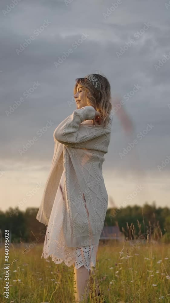 Charming lady putting on long knitted cardigan over dress, then turning around in grassy field against cloudy evening sky in countryside. Vertical shot. Charming lady putting on long knitted cardigan over dress, then turning around in grassy field against cloudy evening sky in countryside. Vertical shot.