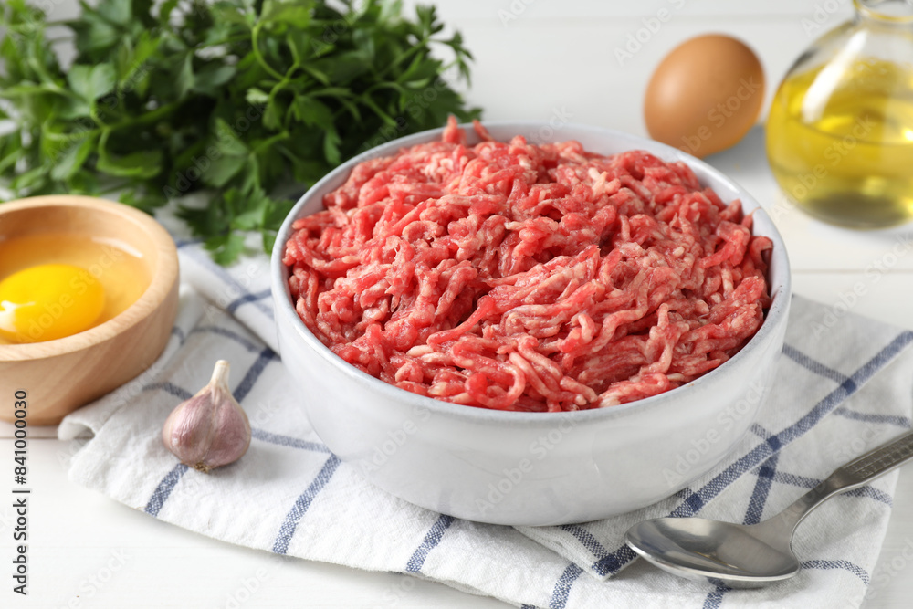 Raw ground meat in bowl, garlic, egg, parsley and spoon on white table, closeup Raw ground meat in bowl, garlic, egg, parsley and spoon on white table, closeup