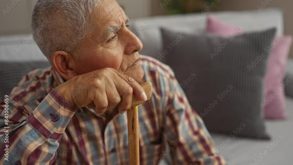 A thoughtful grey-haired hispanic man, dressed in a plaid shirt, rests with a walking stick in a cozy living room with grey and pink cushions. A thoughtful grey-haired hispanic man, dressed in a plaid shirt, rests with a walking stick in a cozy living room with grey and pink cushions.