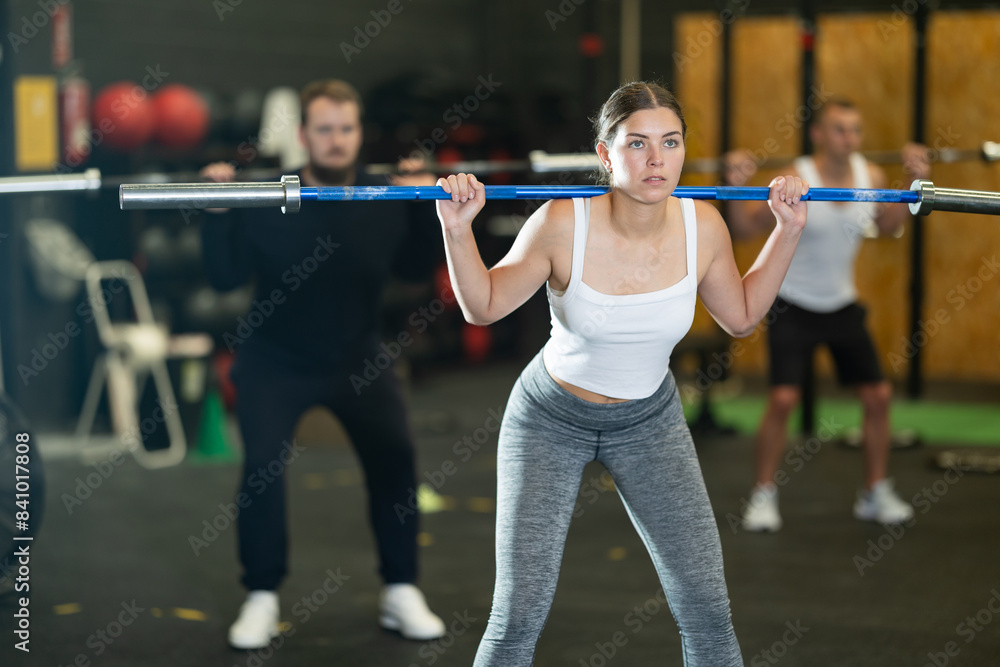 Fototapeta premium Focused motivated sporty young girl performing set of exercises with barbell during intense group workout. Concept of weight training ....