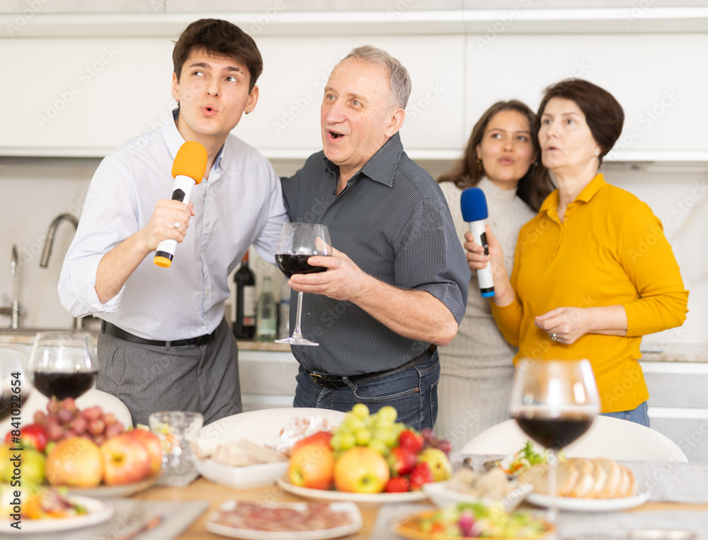 Happy family singing karaoke using microphone during family holiday, parents and adult children at festive table in light kitchen