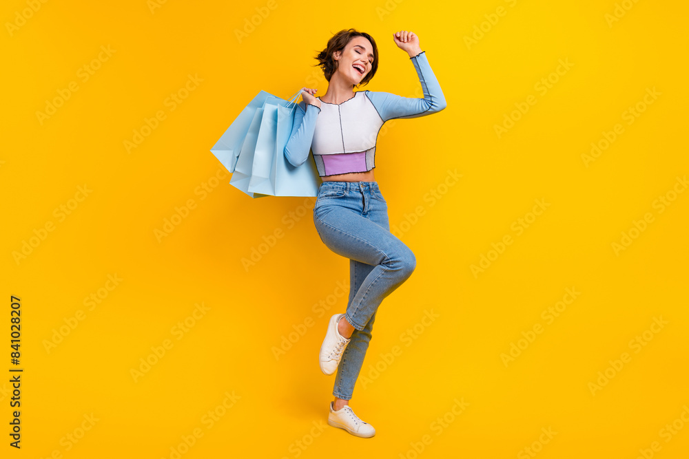 Full body photo of delighted cheerful lady hold mall shop bags raise fist success isolated on yellow color background Full body photo of delighted cheerful lady hold mall shop bags raise fist success isolated on yellow color background