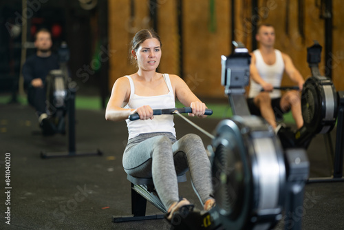 Portrait of concentrated young girl doing intense full body workout in gym, exercising on rowing machine.. © JackF