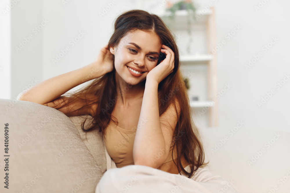 Happy Woman Sitting on a Cosy White Sofa in a Modern Living Room, Looking at Camera with a Confident and Cheerful Smile Happy Woman Sitting on a Cosy White Sofa in a Modern Living Room, Looking at Camera with a Confident and Cheerful Smile