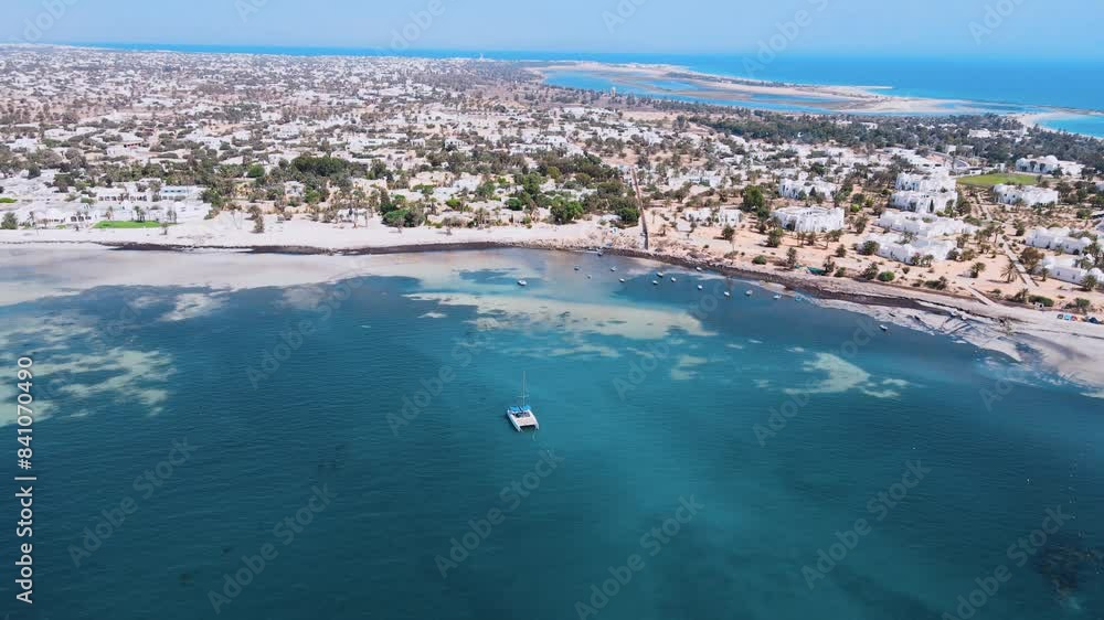 An aerial view of a sailboat is floating on a large body of water, surrounded by a beautiful sandy shores. beach with palm trees and houses nearby. at Tunisia Djerba An aerial view of a sailboat is floating on a large body of water, surrounded by a beautiful sandy shores. beach with palm trees and houses nearby. at Tunisia Djerba