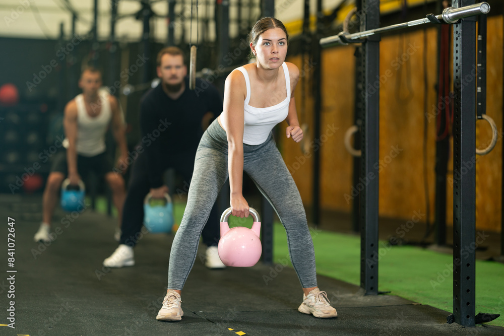 Naklejka premium Sportive young woman lifting heavy kettlebell in sports hall during crossfit workout